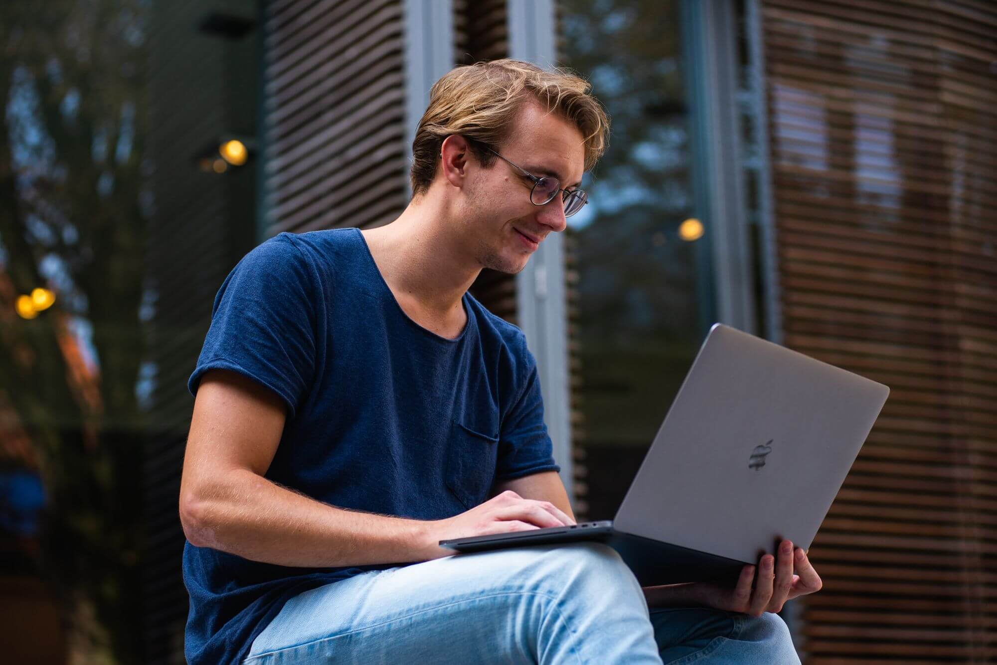 A young man with glasses looks at his laptop outside.